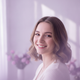 Portrait of a smiling young woman with warm eyes, against a soft background in dusty lilac and bright white.