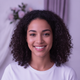 Portrait of a smiling young adult woman with dark curly hair and a natural expression against a soft, light purple background